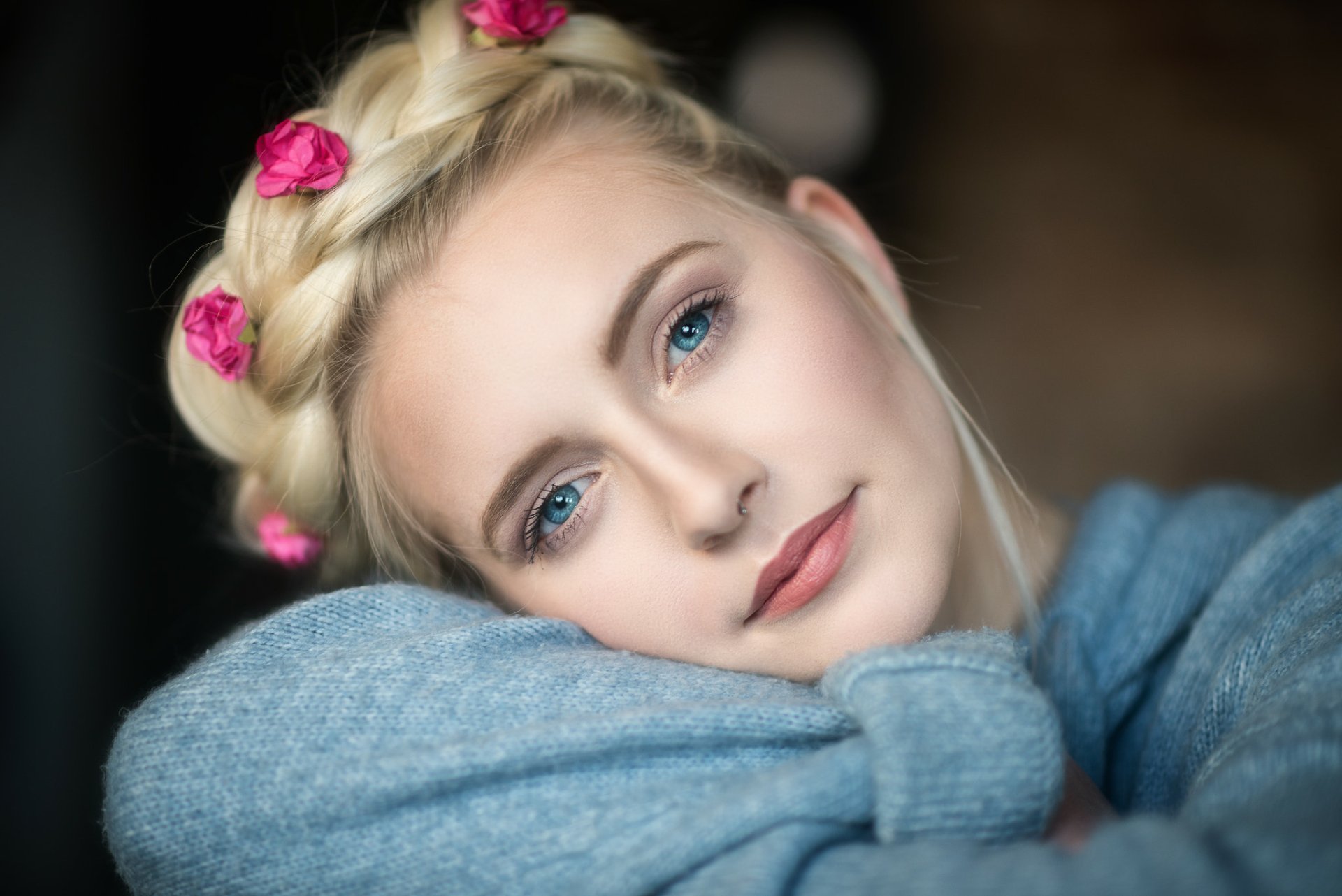 HD desktop wallpaper of a blonde woman with blue eyes, a braided hairstyle adorned with pink flowers, smiling softly and resting her face on her crossed arms.