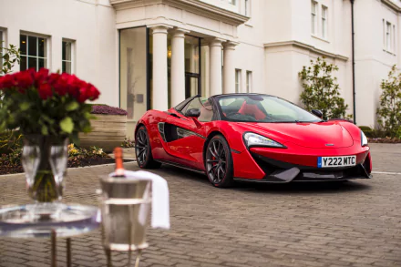 A stunning HD wallpaper featuring a red McLaren 570S Spider supercar parked in front of an elegant building, with roses and a champagne bucket on a table, evoking a luxurious Valentine's Day ambiance.