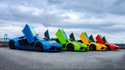 A vibrant HD desktop wallpaper featuring a lineup of Lamborghini Aventadors in blue, green, yellow, and orange, with their doors stylishly open against a cloudy sky backdrop.