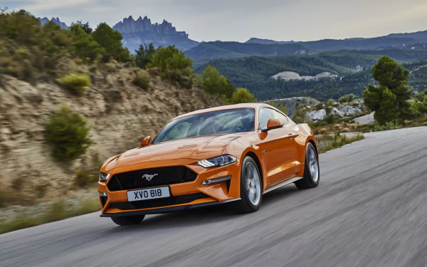 Bright orange Ford Mustang GT muscle car driving on a winding mountain road with rocky hills and greenery in the background, captured in high-definition.