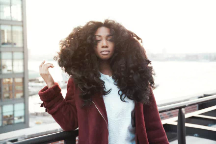 HD desktop wallpaper featuring SZA with voluminous hair, standing on a balcony with a cityscape background.