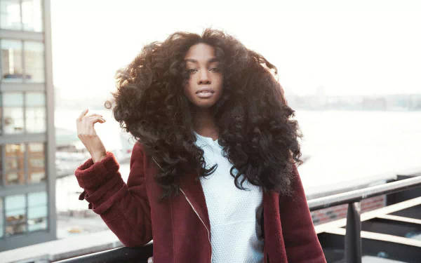 HD desktop wallpaper featuring SZA with voluminous hair, standing on a balcony with a cityscape background.