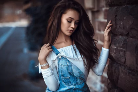 Brunette model with long hair leans against a stone wall, moody expression, shallow depth of field — HD PC desktop wallpaper of a young woman in denim overalls.