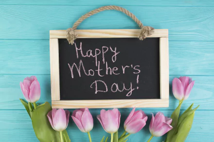 Pink tulip flowers surround a small chalkboard with Happy Mother's Day! written, set against a blue wooden background in a 4K Ultra HD holiday-themed wallpaper.