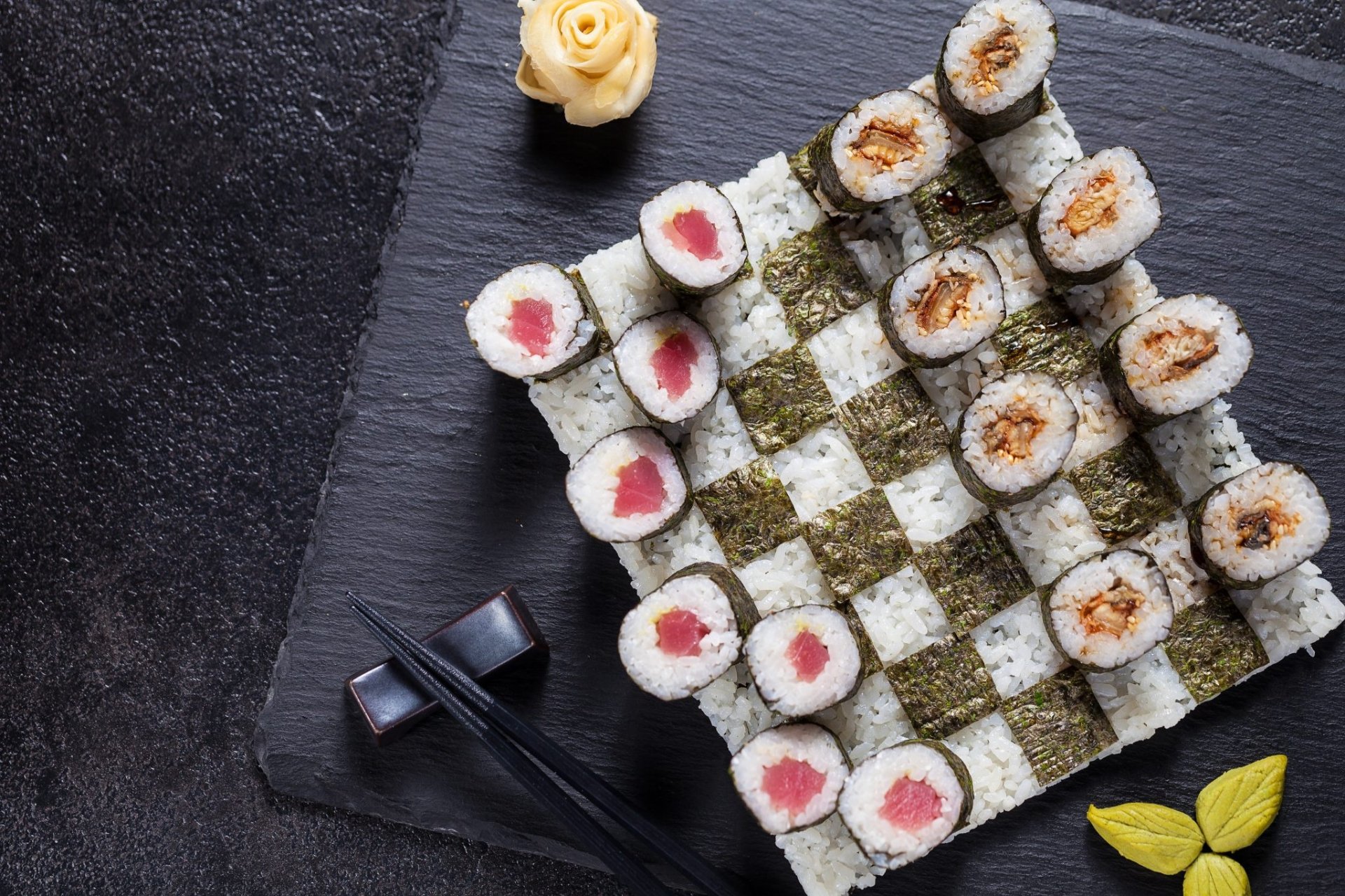 HD desktop wallpaper featuring a still life of sushi arranged on a checkerboard of rice and seaweed, accompanied by chopsticks and garnishes.