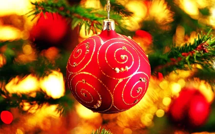 A vibrant red Christmas ornament with gold swirls hangs from a pine branch, surrounded by a warm, glowing holiday background, creating a festive and joyful atmosphere.