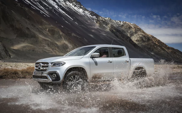 Silver Mercedes‑Benz X‑Class pickup vehicle splashes through a mountain stream against rugged peaks; 4K Ultra HD PC desktop wallpaper background.