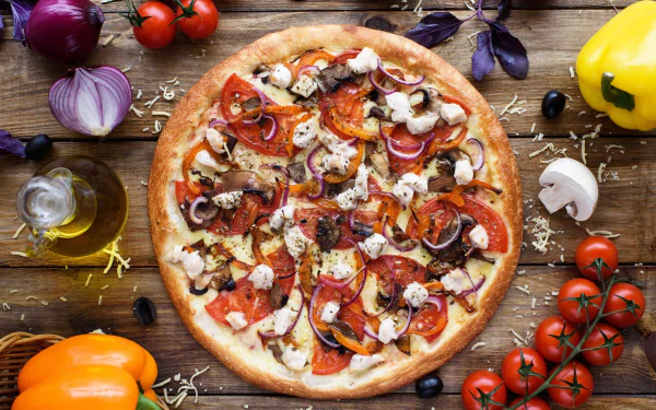 HD desktop wallpaper featuring a still life of a pizza topped with tomatoes, mushrooms, onions, and olives, surrounded by fresh vegetables and herbs on a wooden table.