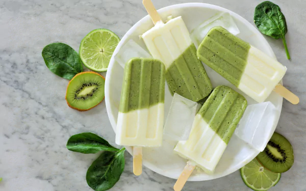 A 4K Ultra HD still life of kiwi and cream popsicles on a white plate, surrounded by fresh kiwi slices, lime halves, and spinach leaves on a marble surface.