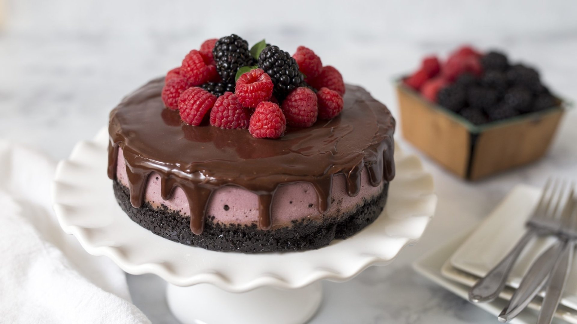 HD desktop wallpaper of a chocolate pastry topped with fresh blackberries and raspberries on a white cake stand, with a box of berries and utensils in the background.
