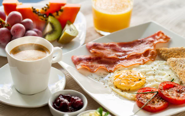 HD desktop wallpaper showing a still life breakfast with egg, bacon, coffee, grilled tomatoes, toast, fresh fruit, and orange juice on a white table.