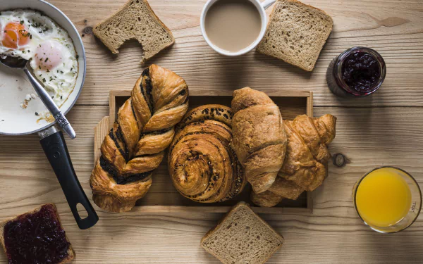 5K Ultra HD PC wallpaper still life of breakfast viennoiserie: croissants, braided and cinnamon rolls on a tray, toast, coffee, orange juice and jam on a wooden table.