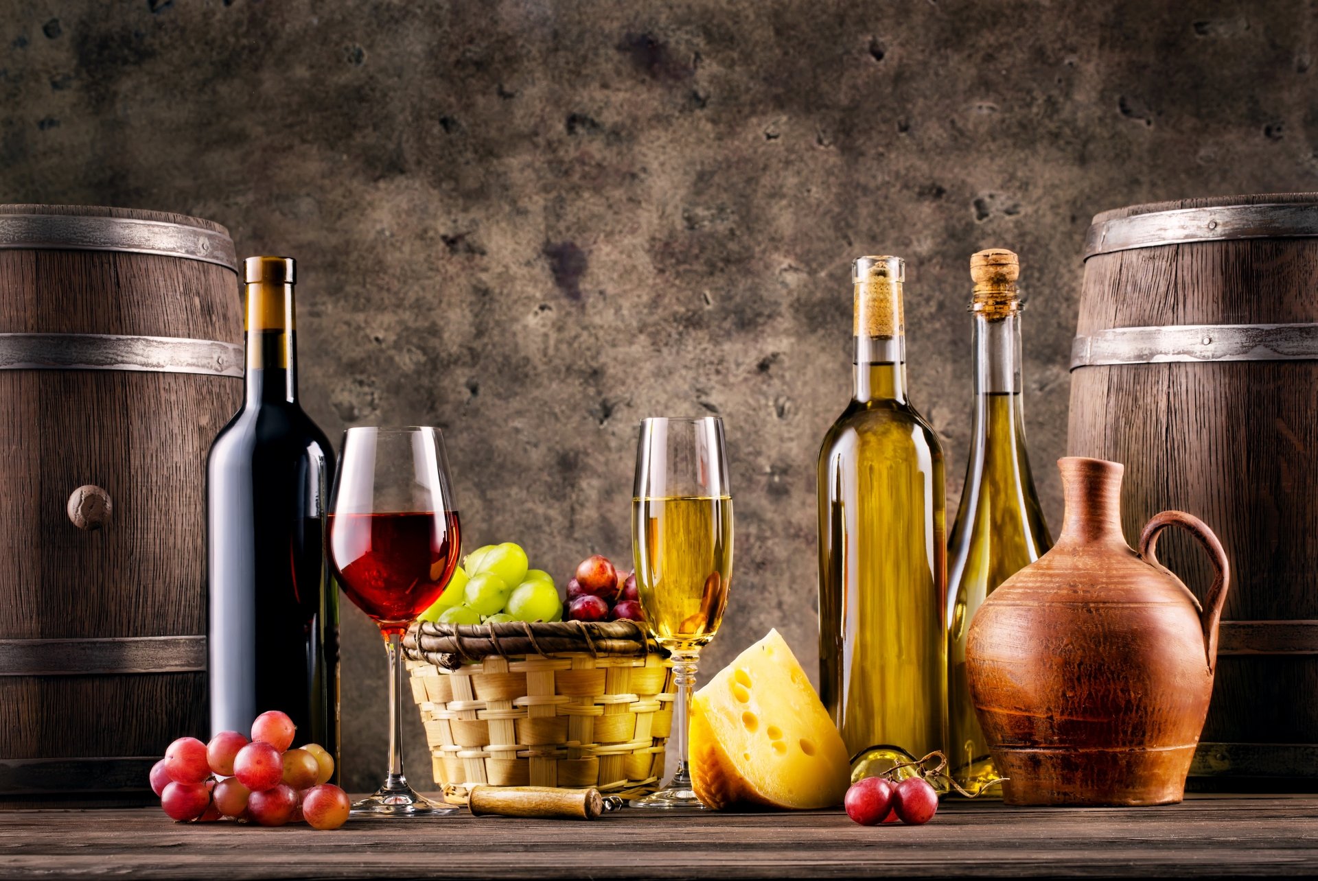 5K Ultra HD PC desktop wallpaper still life: wine bottles and glasses, oil jug, basket of grapes and cheese arranged between wooden barrels.