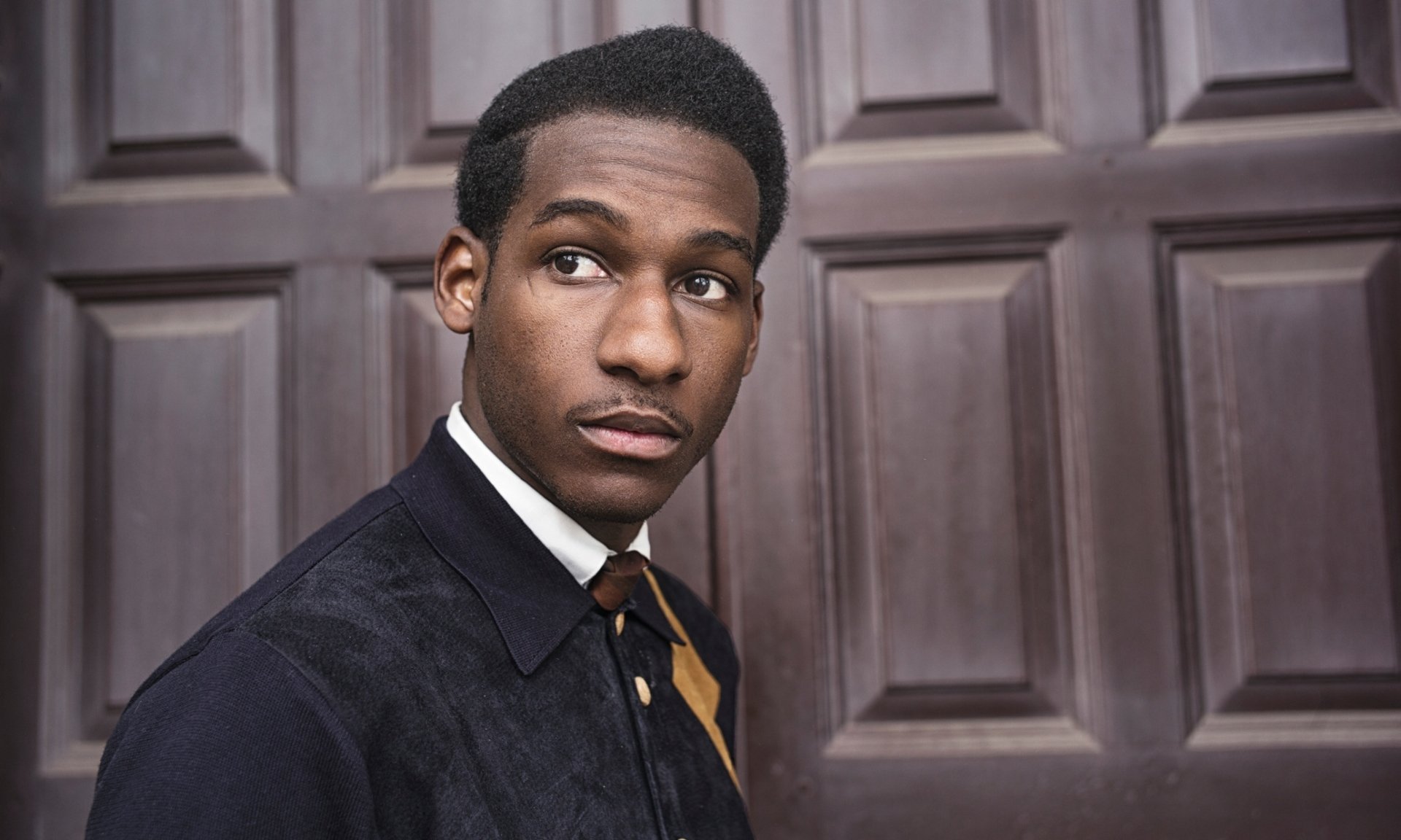 HD desktop wallpaper featuring a portrait of a man dressed in a smart outfit posing in front of a wooden panel door, tagged Leon Bridges.