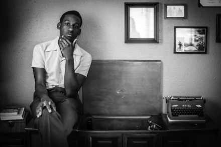 Black and white HD wallpaper of a man sitting thoughtfully beside a suitcase with a vintage typewriter, framed photos in the background.