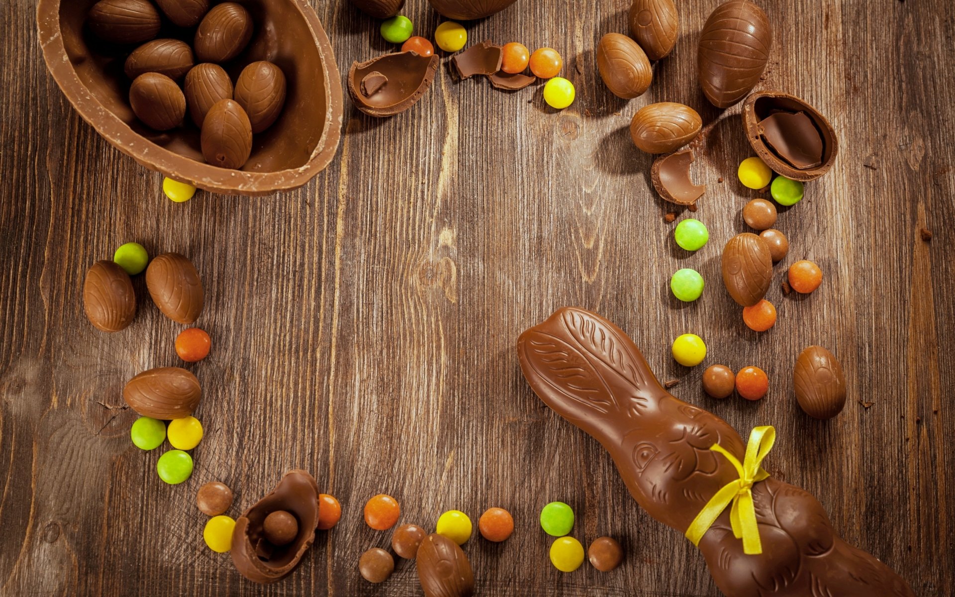 2K Quad HD PC desktop wallpaper: chocolate Easter eggs, colorful candy and a ribbon-tied chocolate bunny arranged on a rustic wooden surface — holiday Easter scene.