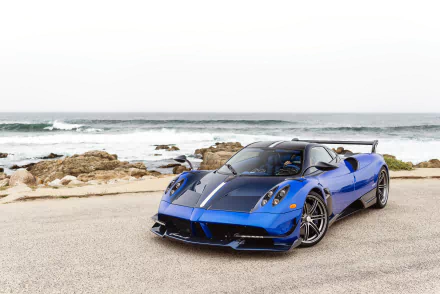 Blue Pagani Huayra Roadster BC supercar parked by the rocky ocean shore under a clear sky, captured in high-definition for a PC desktop wallpaper.