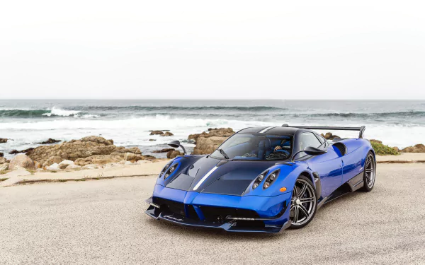 Blue Pagani Huayra Roadster BC supercar parked by the rocky ocean shore under a clear sky, captured in high-definition for a PC desktop wallpaper.