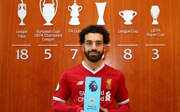 HD desktop wallpaper of Egyptian soccer star Mohamed Salah in a Liverpool jersey, holding the Premier League Player of the Month award with his major trophies displayed behind him.