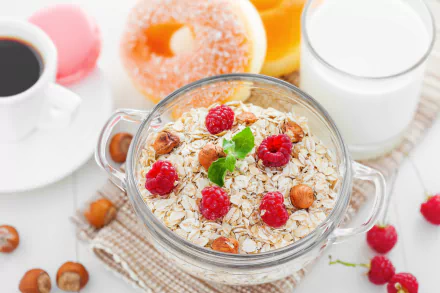 A 4K Ultra HD image of a breakfast scene featuring a glass bowl of muesli with raspberries and milk, accompanied by coffee, a donut, and a glass of milk on a white table.