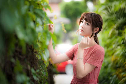 A brunette Asian woman in a pink dress poses thoughtfully amid lush greenery, captured with a shallow depth of field in this 4K Ultra HD desktop wallpaper.