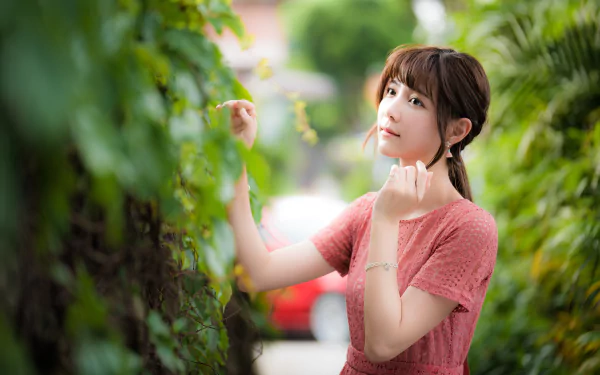 A brunette Asian woman in a pink dress poses thoughtfully amid lush greenery, captured with a shallow depth of field in this 4K Ultra HD desktop wallpaper.