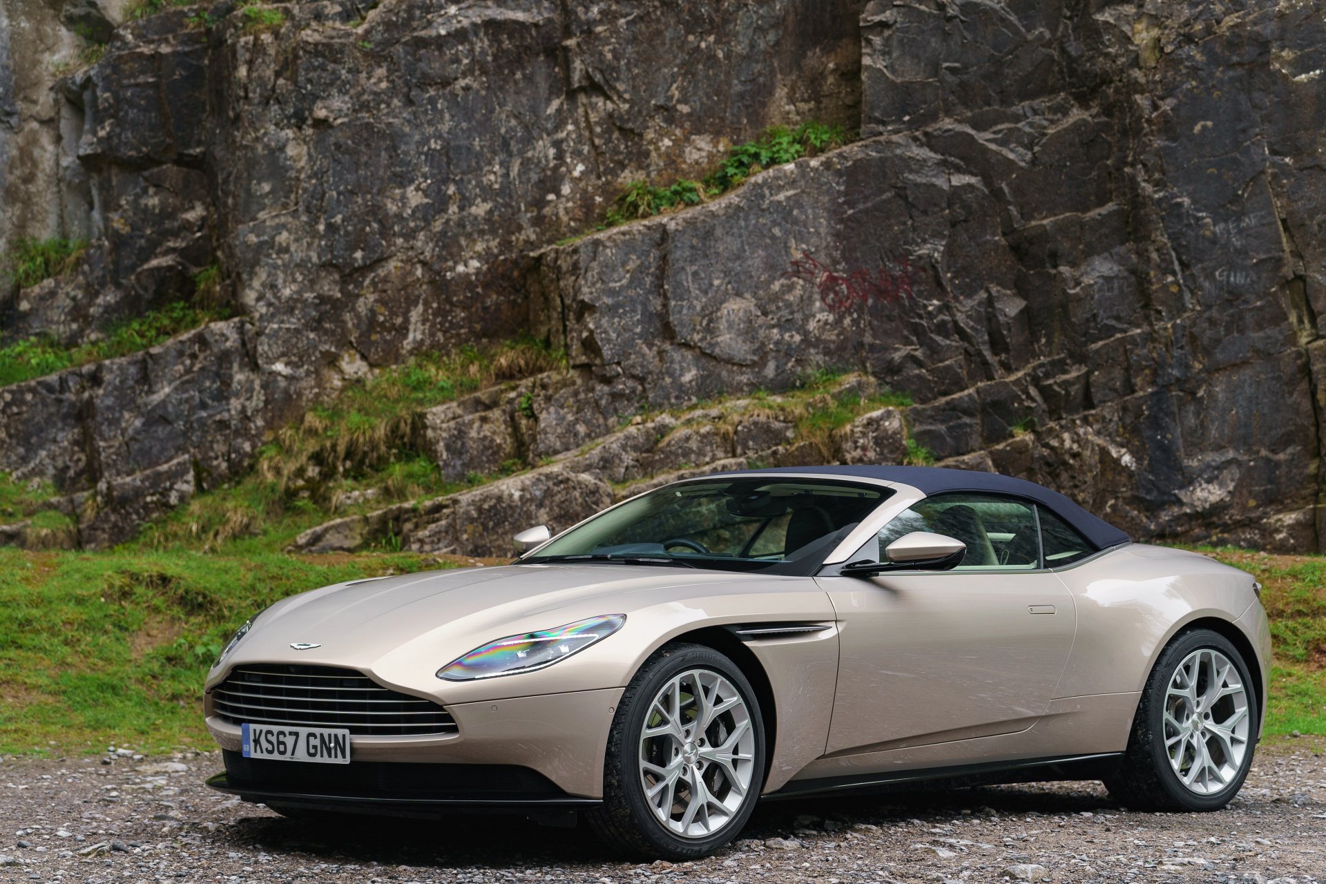 Silver Aston Martin DB11 cabriolet grand tourer parked on a gravel surface with rocky cliffs in the background, shown in 4K Ultra HD quality.