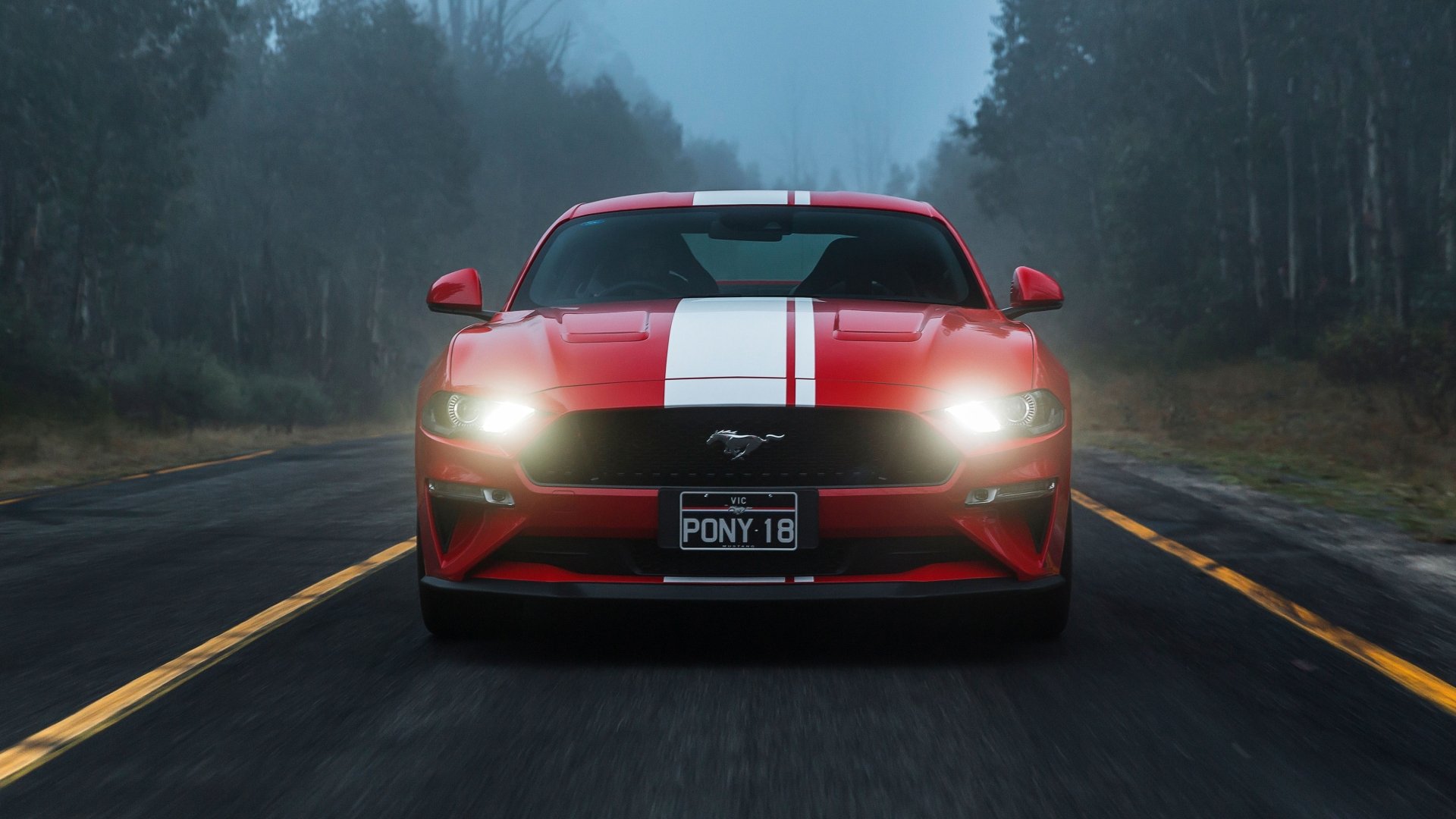 Red Ford Mustang GT muscle car with white racing stripe, headlights on, front view on a foggy road - 4K Ultra HD PC desktop wallpaper and background.