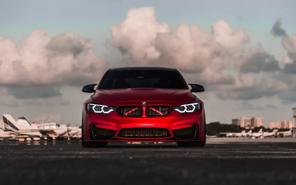 HD desktop wallpaper featuring a red BMW M4 car, showcasing its aggressive front design and illuminated headlights against a cloudy sky background.