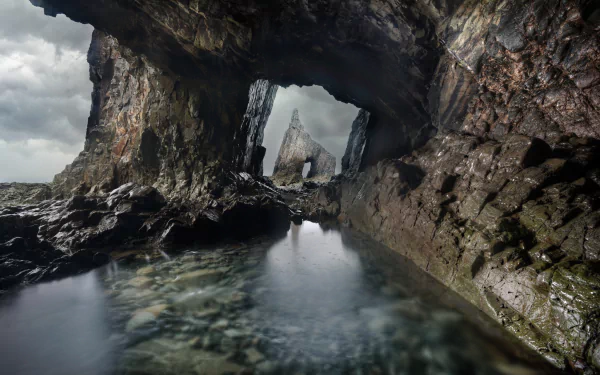 HD PC desktop wallpaper background: dramatic coastal rock arch forming a cavernous tunnel over a reflective tidal pool, with a distant sea stack and moody cloudy sky.