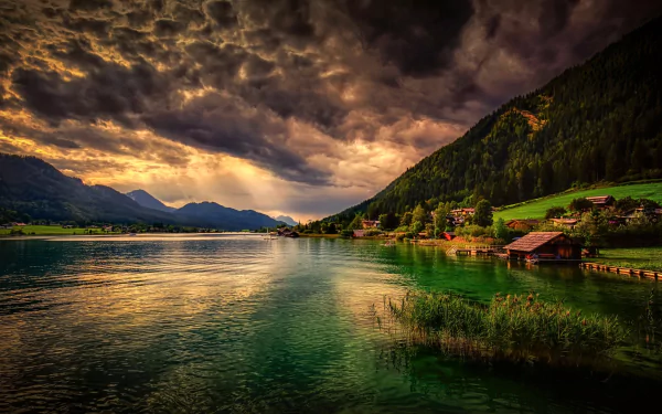 HD desktop wallpaper: A serene village by a tranquil lake, surrounded by majestic mountains under a dramatic cloudy sky, featuring a house and lush green landscape.