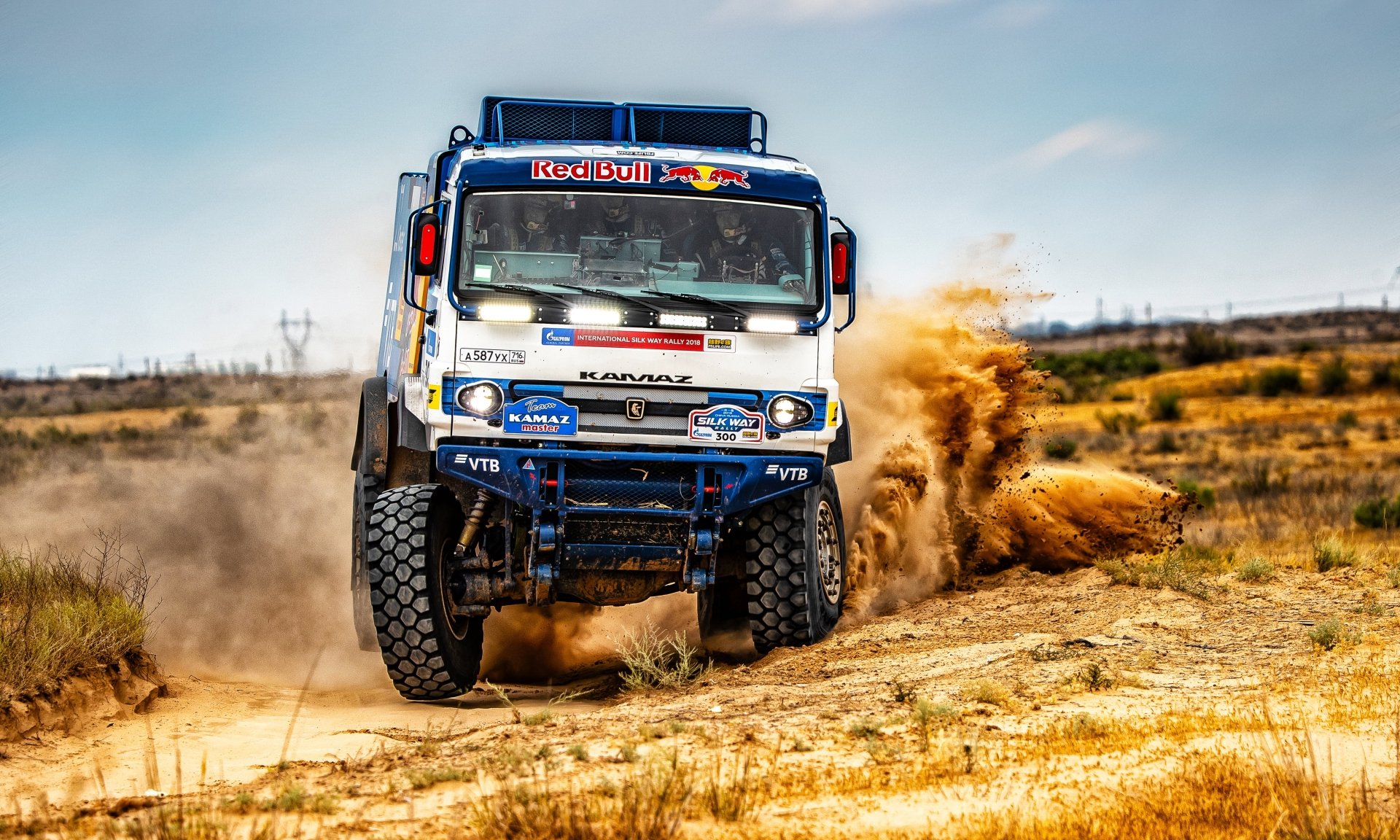 A Red Bull Kamaz rally truck speeds through sandy terrain, kicking up dust under a clear sky in this 4K Ultra HD sports vehicle wallpaper.
