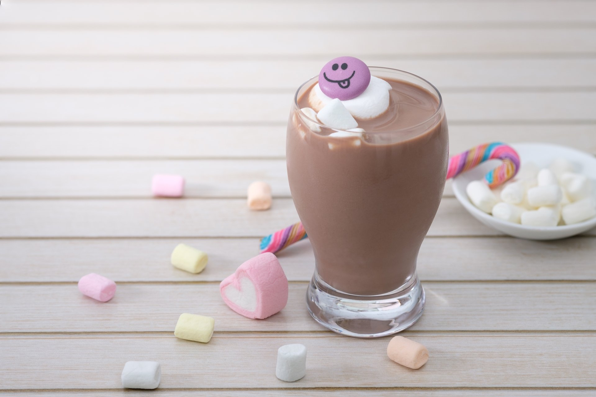 A 4K Ultra HD still life of a glass of hot chocolate topped with marshmallows and a smiling pink candy, surrounded by pastel marshmallows and a candy cane on a wooden surface.