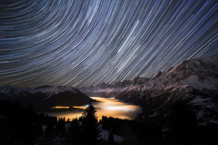 HD desktop wallpaper of a mountain landscape at night, featuring swirling star trails above snowy peaks and a glowing valley blanketed in mist.