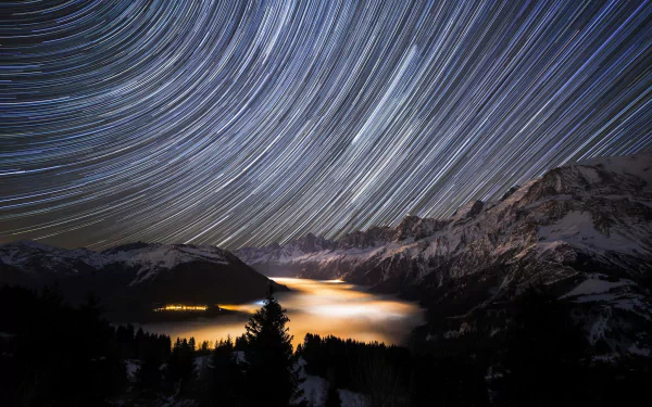 HD desktop wallpaper of a mountain landscape at night, featuring swirling star trails above snowy peaks and a glowing valley blanketed in mist.