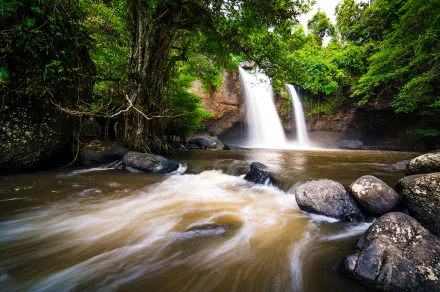 HD desktop wallpaper capturing a serene forest river flowing over a twin waterfall surrounded by lush green trees and smooth rocks.