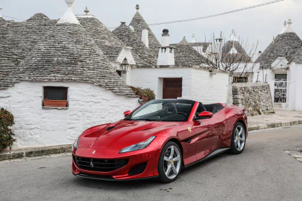 Red Ferrari Portofino supercar parked on a street with traditional white stone houses and conical roofs, captured in stunning 4K Ultra HD for PC desktop wallpaper.