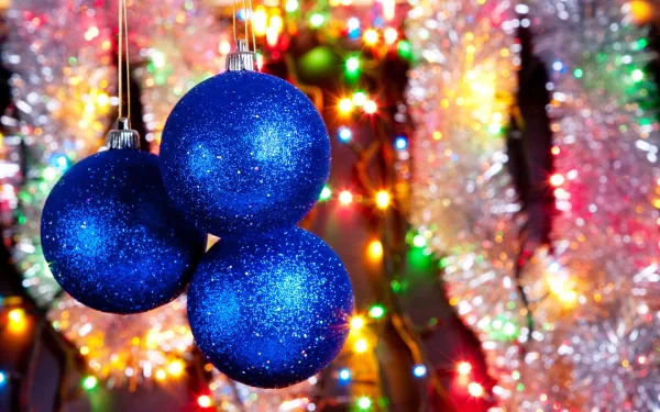 HD desktop wallpaper featuring three blue glittery Christmas ornaments against a vibrant background of colorful holiday lights and tinsel.