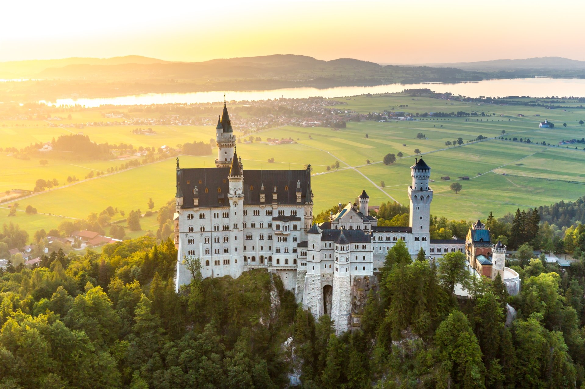 A stunning 4K Ultra HD landscape of Germany's man-made Neuschwanstein Castle surrounded by lush greenery and distant hills at sunrise.