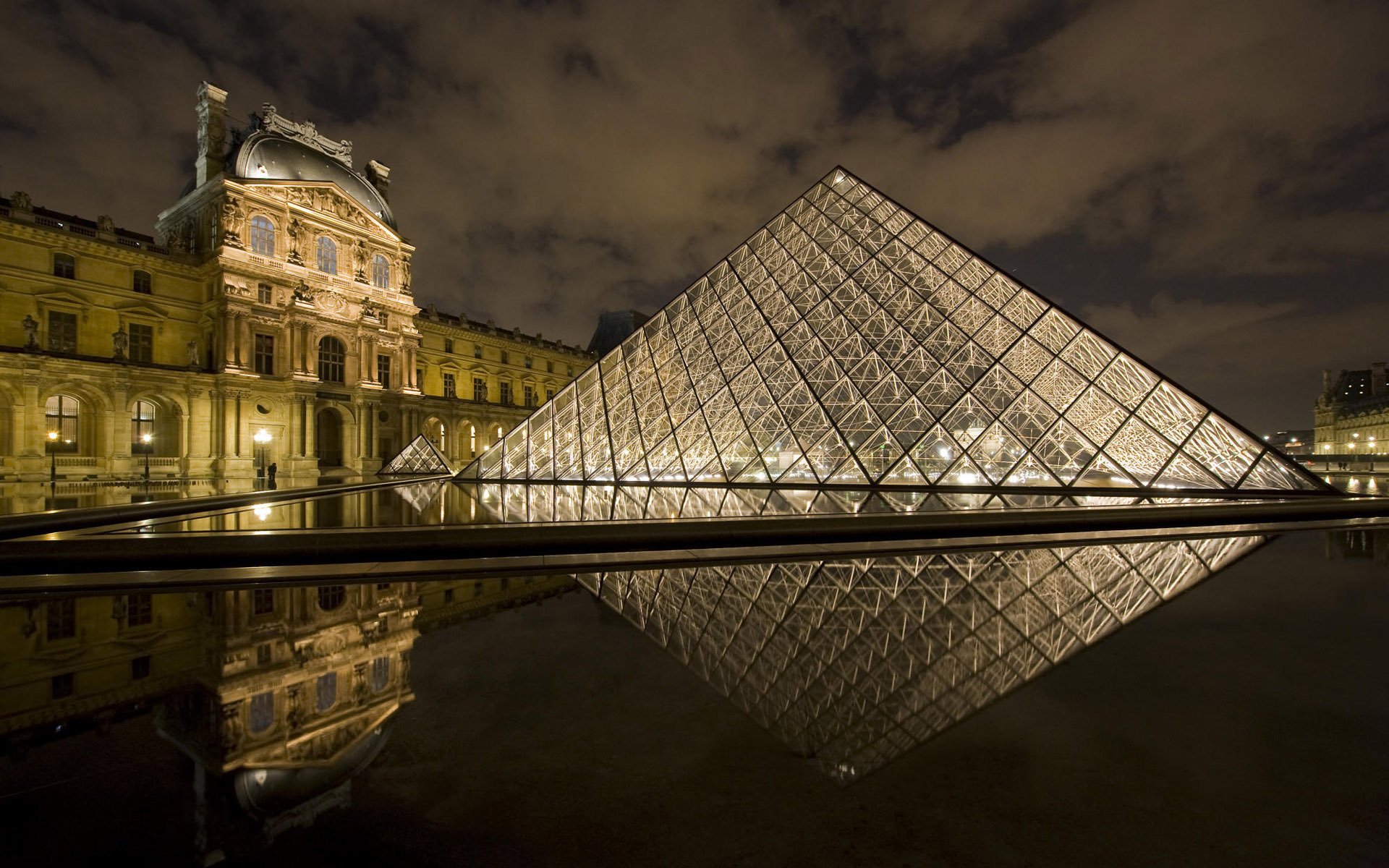 The Louvre in Paris at night, showcasing the iconic glass pyramid reflecting in the water, with the historic museum illuminated against a dramatic sky. HD desktop wallpaper.