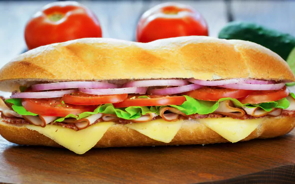 Food, sandwich 5K Ultra HD PC desktop wallpaper: close-up of a loaded deli baguette with ham, cheese, lettuce, tomato and red onion on a wooden board.