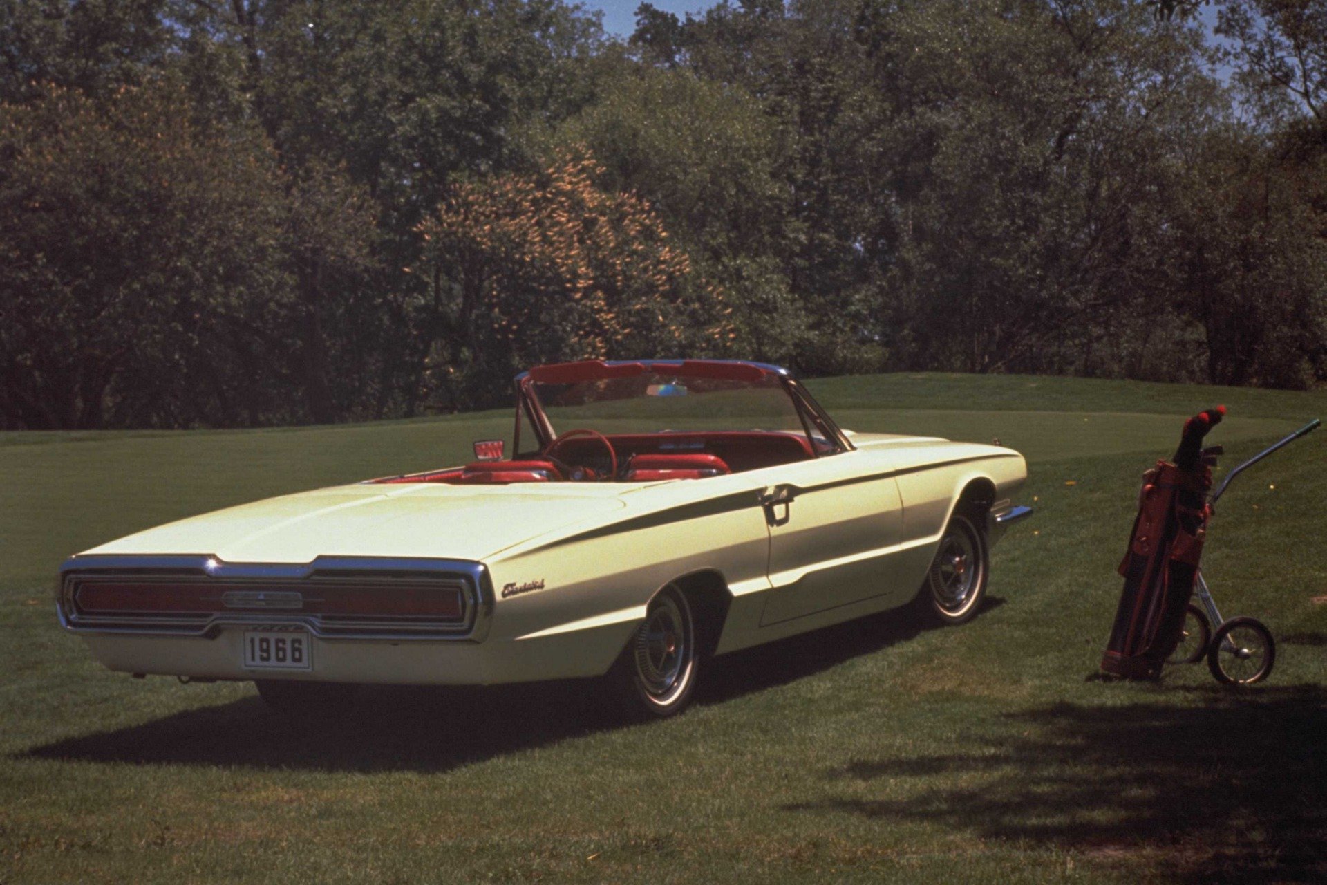 HD desktop wallpaper featuring a vintage Ford Thunderbird convertible parked on a grassy lawn with golf clubs nearby and trees in the background.