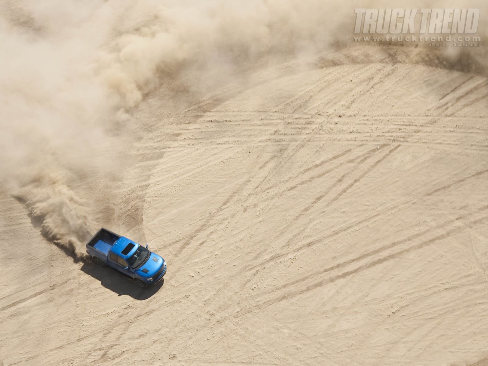 HD PC desktop wallpaper featuring a blue Ford Raptor creating tire marks and kicking up dust on a sandy terrain.