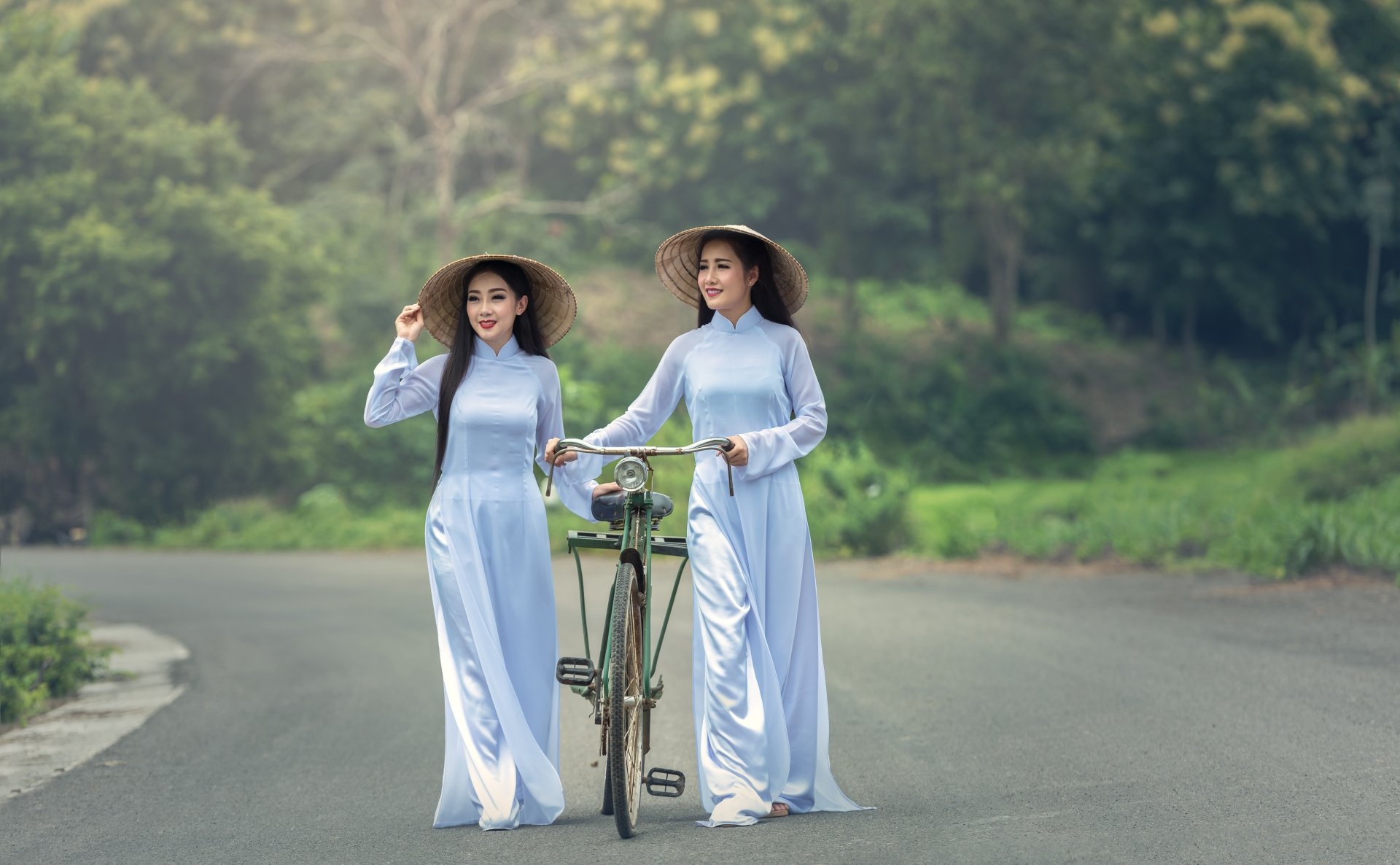 Two Asian women with black long hair smile wearing traditional dresses and conical hats, standing on a road with a bicycle, captured in 4K Ultra HD.