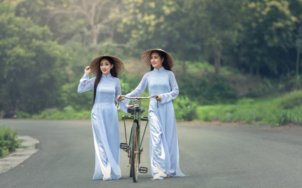 Two Asian women with black long hair smile wearing traditional dresses and conical hats, standing on a road with a bicycle, captured in 4K Ultra HD.