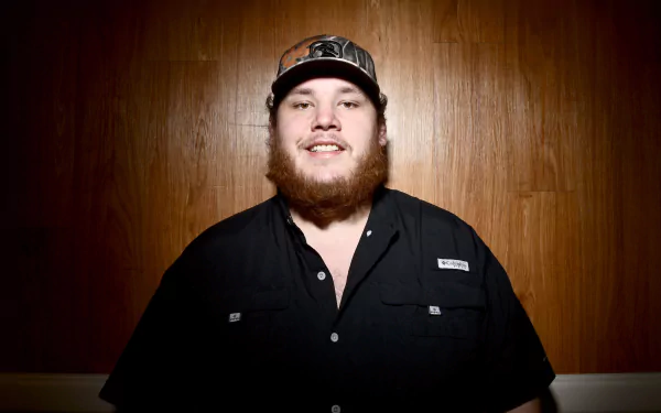 HD desktop wallpaper featuring a smiling man in a black shirt and cap against a wooden background.