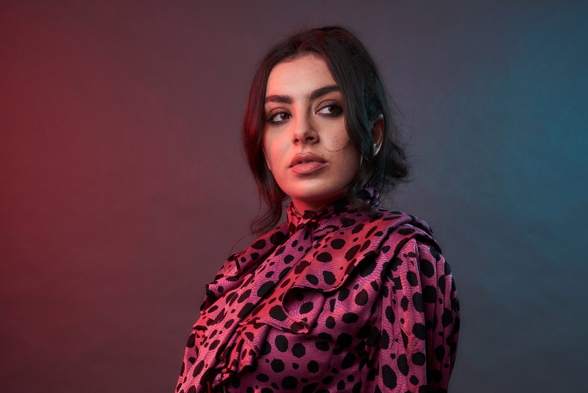 5K desktop wallpaper: close-up of a black-haired woman with brown eyes in a pink polka-dot blouse, red-blue studio lighting, evoking a pop singer/music aesthetic.