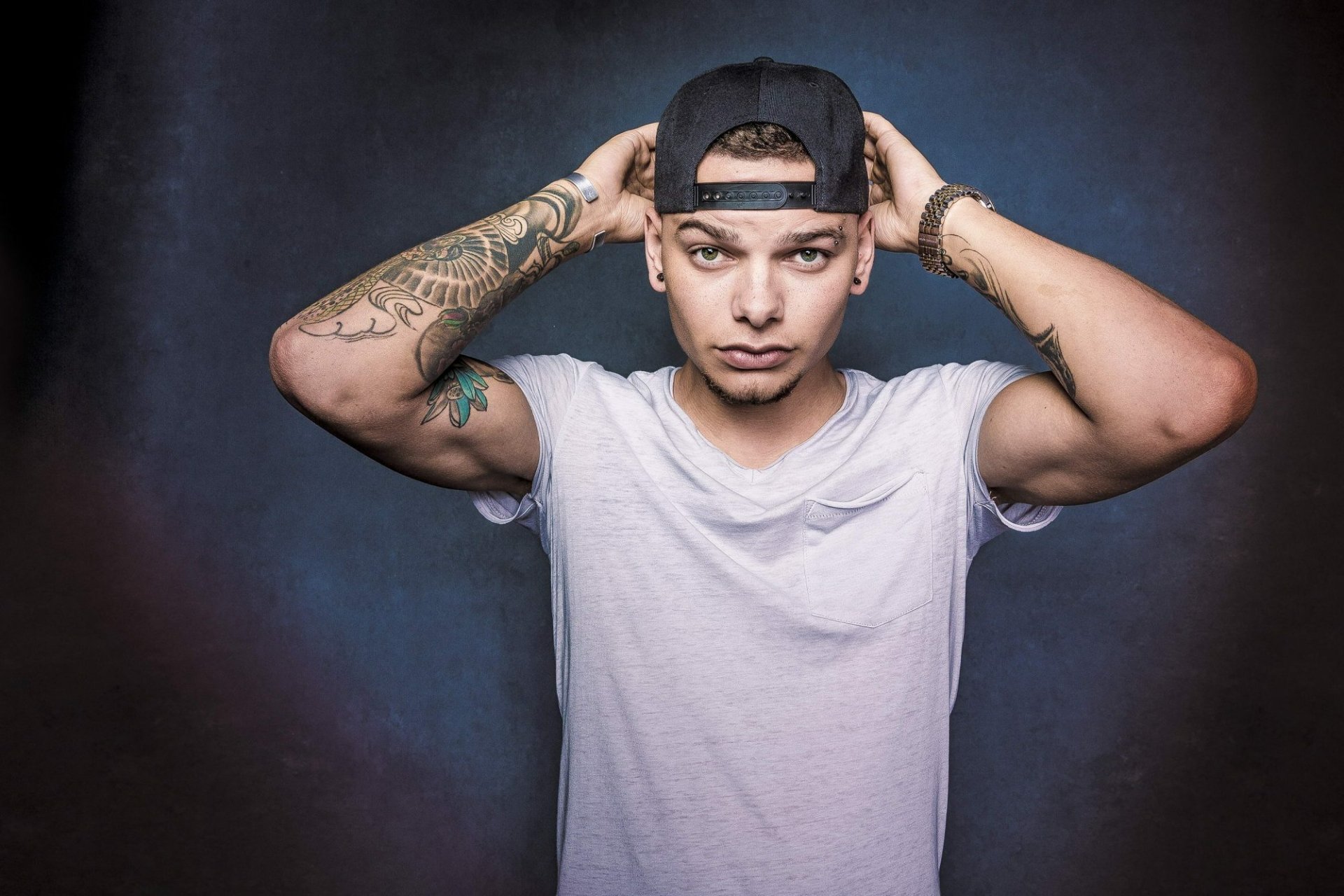 HD desktop wallpaper featuring a poised individual adjusting a baseball cap, with visible tattoos, set against a muted backdrop.