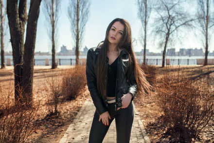 HD desktop wallpaper featuring a brunette model with long hair wearing a jacket, standing on a path with trees and cityscape blurred in the background using depth of field.