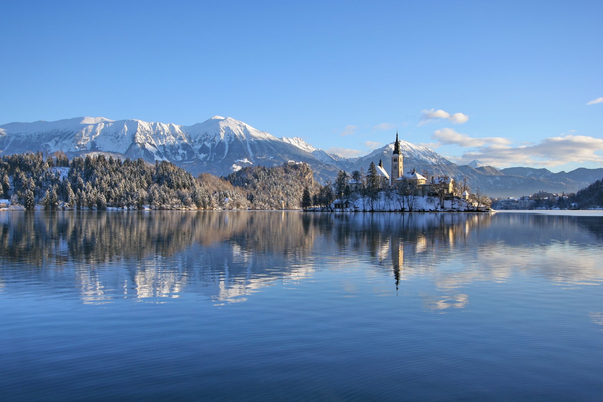 Winter view of the Assumption of Mary Church on a serene lake surrounded by snow-covered trees and mountains, captured in HD PC desktop wallpaper quality.